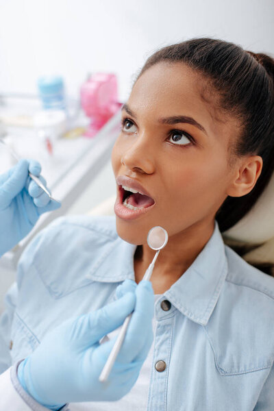 dentist in latex gloves holding dental equipment near african american patient with opened mouth 