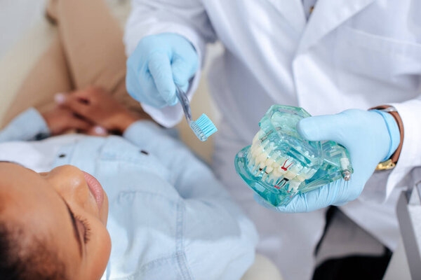 selective focus of dentist in latex gloves holding teeth model and toothbrush near african american patient 