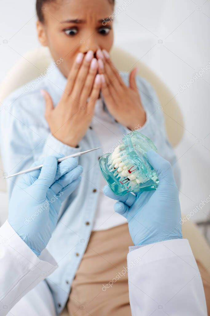 selective focus of dentist in latex gloves holding teeth model and dental instrument near shocked african american woman 