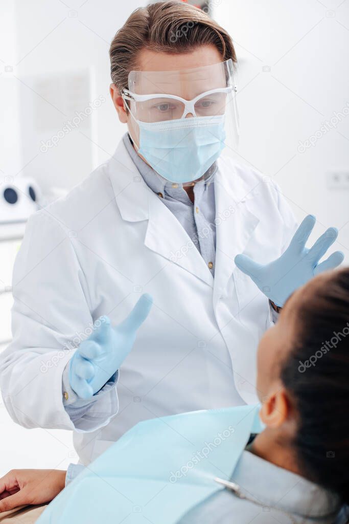 selective focus of dentist in face shield, medical mask and latex gloves gesturing near african american woman 