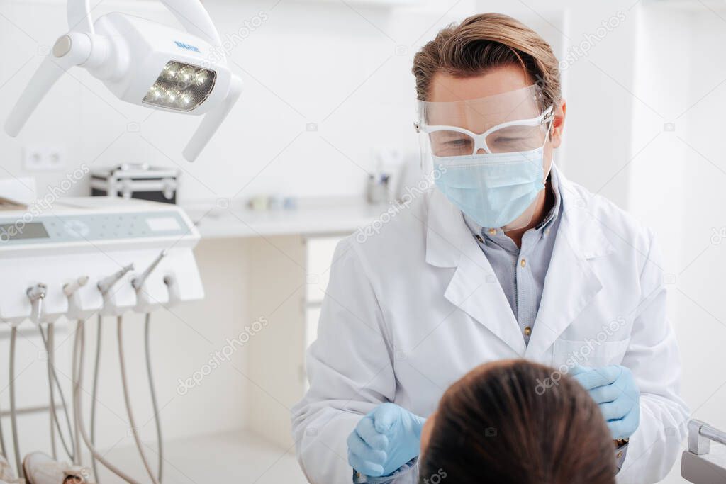 selective focus of dentist in face shield, medical mask and latex gloves near woman 