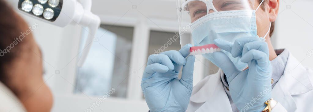panoramic shot of dentist in medical mask, face shield and latex gloves holding dental prosthesis near african american woman 