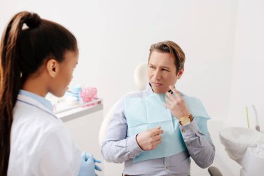 man with tooth pain looking at african american dentist in clinic  clipart