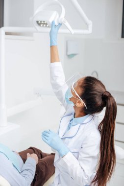Tıbbi maske takmış Afro-Amerikan diş hekimi tıbbi lambaya dokunuyor. 