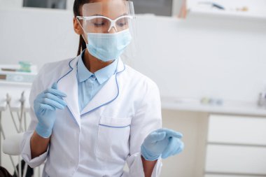 Tıbbi maskeli Afro-Amerikan dişçi, yüz kalkanı ve lateks eldivenler kliniği işaret ediyor. 