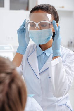 Tıbbi maskeli Afro-Amerikan dişçinin seçici odak noktası hastanın yanındaki yüz kalkanına dokunuyor. 