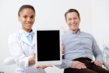 Neşeli Afrika kökenli Amerikalı dişçinin yanındaki mutlu hastanın seçici odağı elinde boş ekranlı dijital tablet tutuyor.