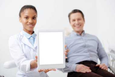 Mutlu bir hastanın yanında beyaz ekranlı dijital tablet tutan neşeli Afro-Amerikan dişçi seçici odağı 