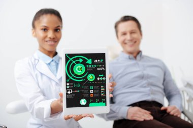 Neşeli Afrikalı Amerikalı dişçinin seçici odak noktası elinde dijital tabletle ekranda mutlu bir hastanın yanında çizelgeler ve grafikler tutması. 