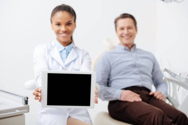 Gülümseyen Afro-Amerikan dişçinin seçici odak noktası mutlu hastanın yanında boş ekranlı dijital tablet tutması. 
