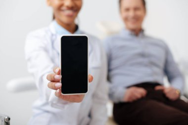 Mutlu bir hastanın yanında beyaz ekran ile akıllı telefonu tutan neşeli Afro-Amerikan dişçi seçici odağı 