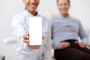 Mutlu bir hastanın yanında beyaz ekranlı akıllı telefon tutan neşeli Afro-Amerikan dişçi seçici odağı 