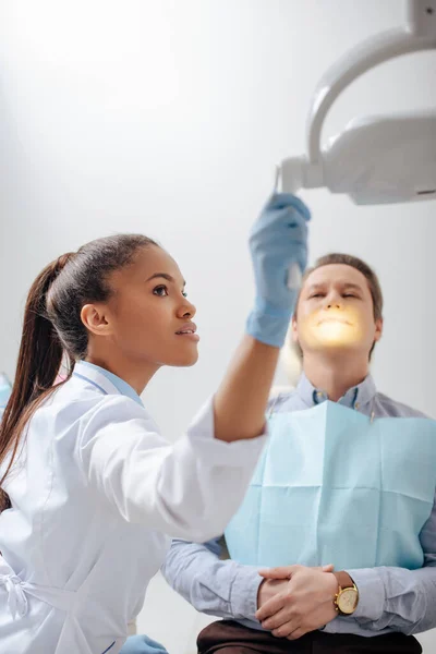 患者の近くの医療ランプに触れるラテックス手袋の美しいアフリカ系アメリカ人歯科医の選択的な焦点 — ストック写真