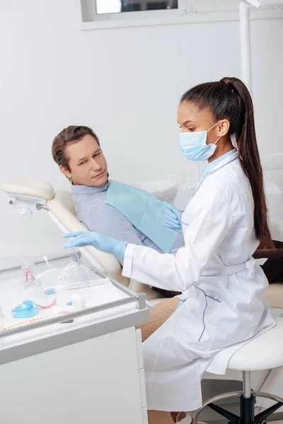 african american dentist in medical mask and man looking at face shield in clinic