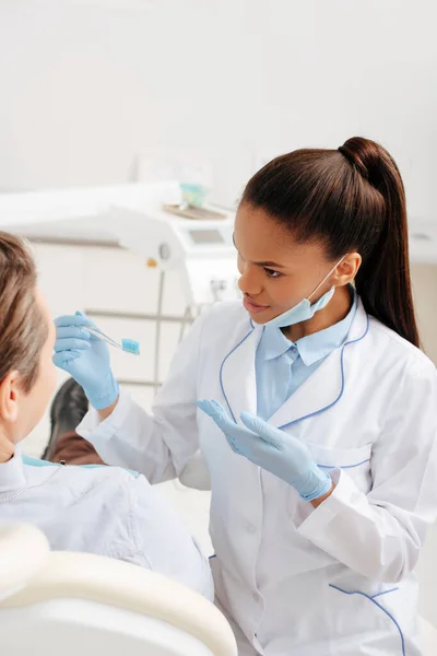 戴乳胶手套的非洲裔美国牙医用手拿着牙刷靠近病人的选择焦点 — 图库照片