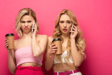 Kağıt bardaklarla kafası karışık sarışın kızlar pembe arka planda akıllı telefonlardan konuşuyorlar.