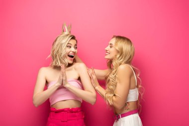 Smiling girl looking at excited blonde friend in crown on pink background clipart