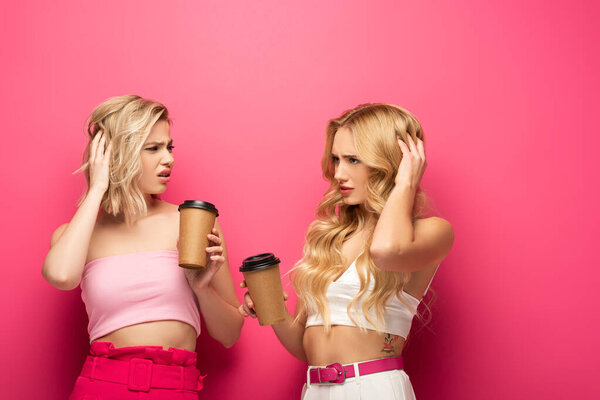Pensive blonde sisters holding coffee to go on pink background