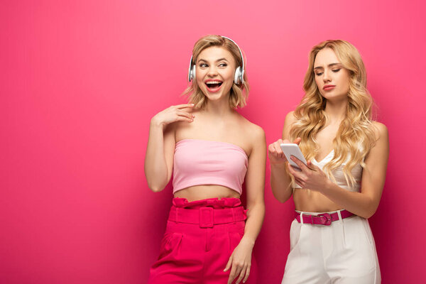 Cheerful blonde girl in headphones near friend using smartphone on pink background