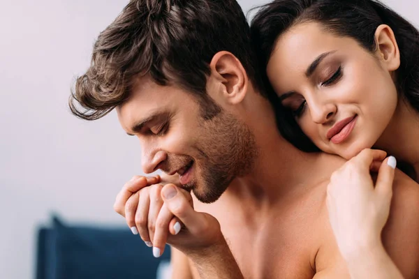 Beautiful woman with closed eyes smiling and hugging man holding female hand in bedroom