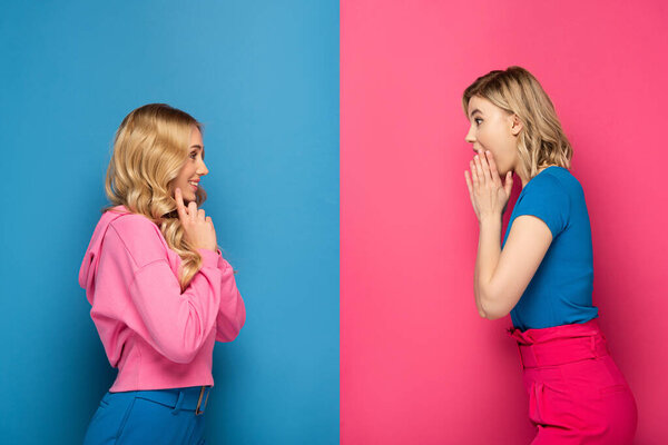 Side view of shocked and smiling blonde sisters looking at each other on pink and blue background