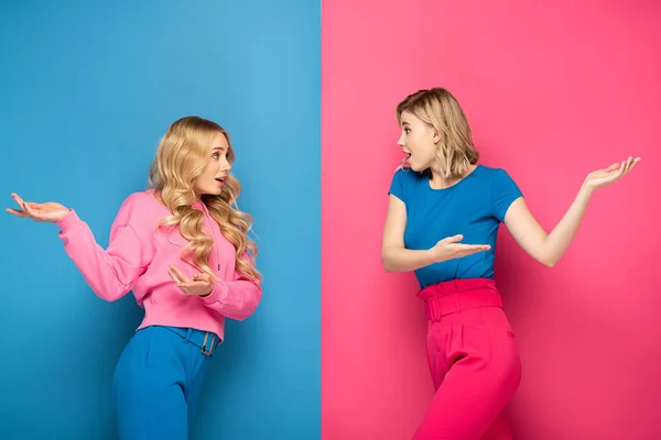 Side view of shocked blonde girls pointing with hands on pink and blue background