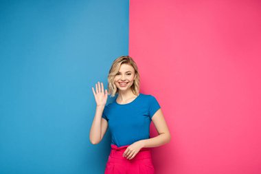Gülümseyen sarışın kadın pembe ve mavi arka planda kameraya el sallıyor.