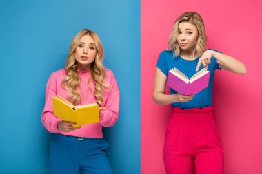 Mavi ve pembe arkaplan üzerine kitaplar tutarken kameraya bakan kafası karışmış sarışın kız kardeşler.