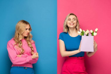 Kızgın sarışın kız pembe ve mavi arka planda buketi olan mutlu kız kardeşine bakıyor.
