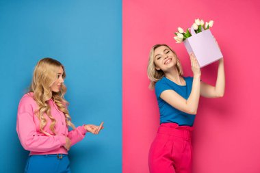 Sarışın kız, pembe ve mavi arka planda buketi olan gülümseyen kız kardeşe orta parmağını gösteriyor.