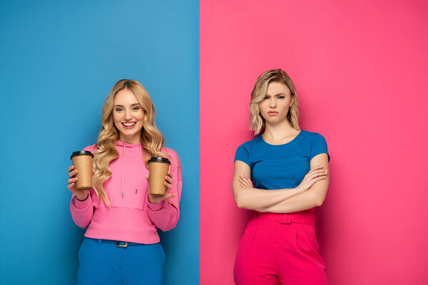 Smiling blonde girl holding paper cups near offended sister on pink and blue background