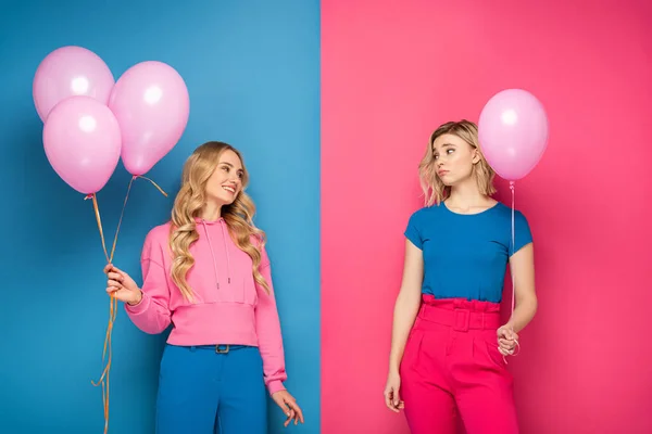 Menina Sorridente Olhando Chateado Irmã Loira Com Balão Fundo Azul — Fotografia de Stock