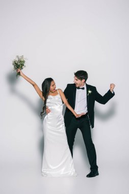 full length view of happy, elegant interracial newlyweds dancing on white background clipart