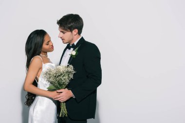 Yakışıklı damat ve çekici Afro-Amerikan gelini düğün buketi ile beyaz arka planda yüz yüze duruyorlar.