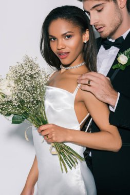 Güzel Afro-Amerikan kızı düğün buketi ile kameraya bakarken damat beyaz arka planda omzuna dokunuyor.