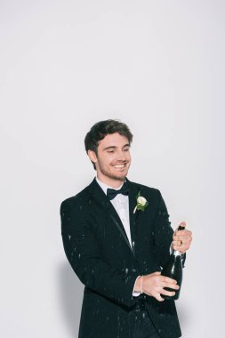 cheerful bridegroom opening bottle near splashes of champagne on white background clipart