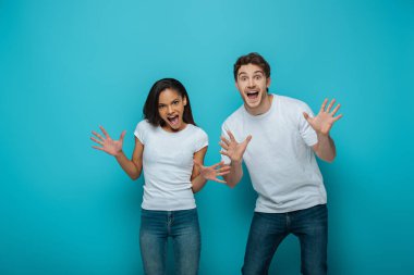 cheerful interracial couple showing frightening gestures at camera on blue background clipart