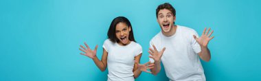 panoramic shot of cheerful interracial couple showing frightening gestures at camera on blue background clipart