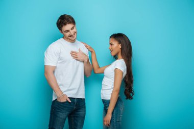 shy young man holding hand on chest while african american girl touching his shoulder on blue background clipart