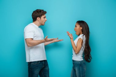 side view of young interracial couple quarreling and gesturing on blue background clipart