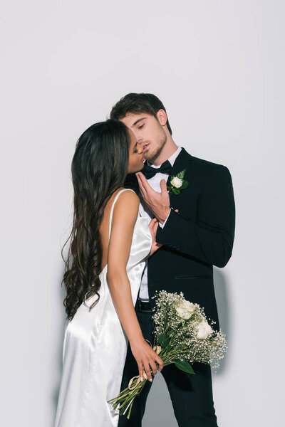 young bridegroom touching face and kissing beautiful african american bride on white background