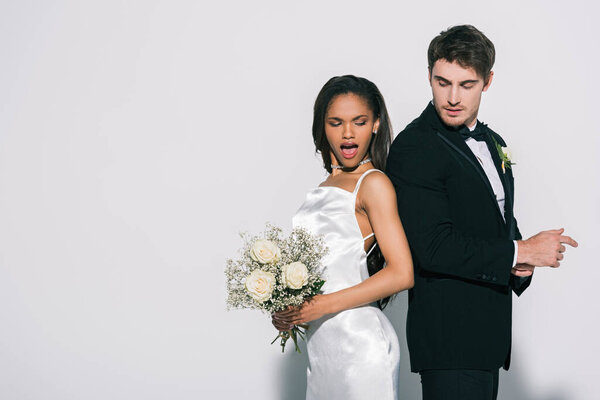 handsome bridegroom and attractive african american bride standing back to back on white background