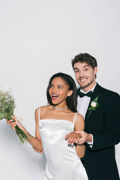 cheerful bridegroom showing wedding on hand of beautiful african american bride on white background