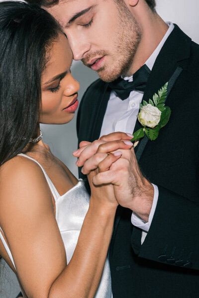 young interracial newlyweds holding hands while standing face to face with closed eyes on white background