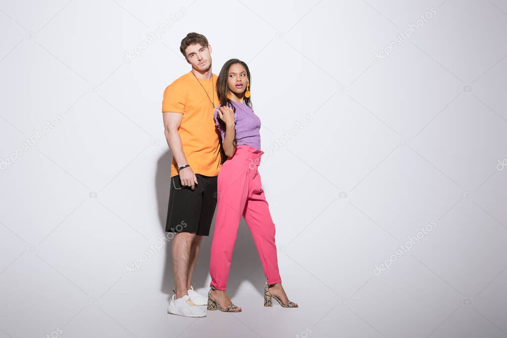 full length view of stylish interracial couple in bright clothes posing on white background