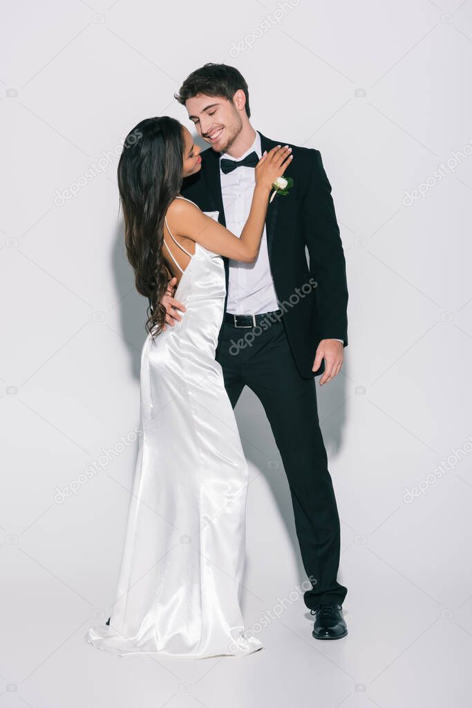 full length view of african american bride in satin wedding dress touching happy bridegroom on white background