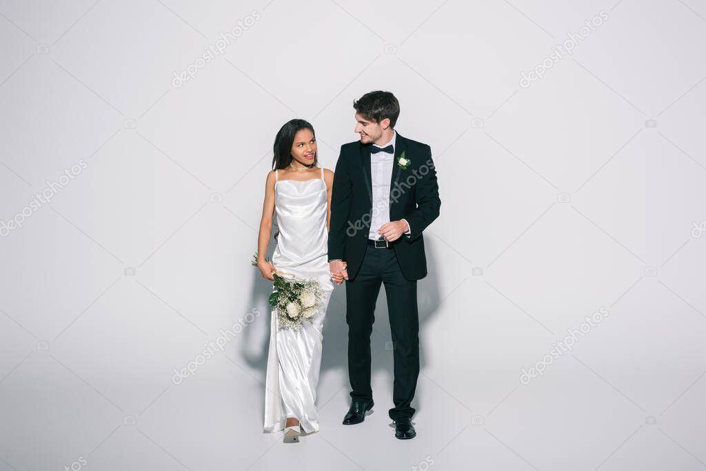 full length view of happy interracial newlyweds holding hands and looking at each other while walking on white background
