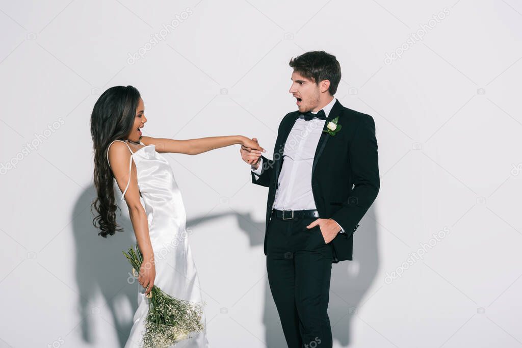 excited bridegroom holding hand of happy african american bride on white background
