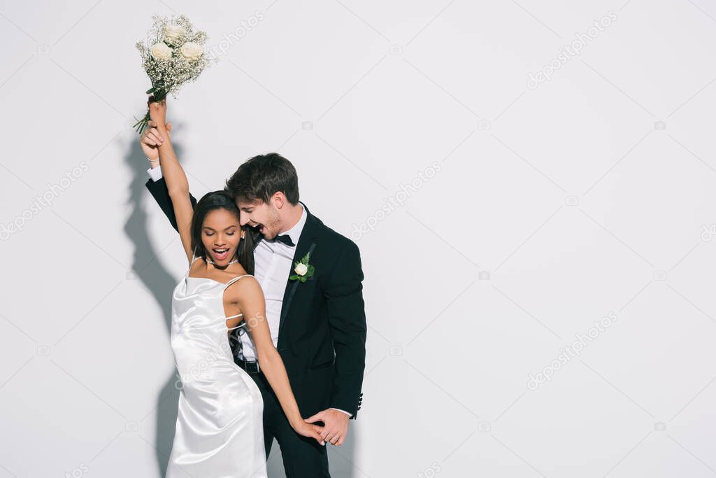 happy, elegant interracial newlyweds dancing on white background