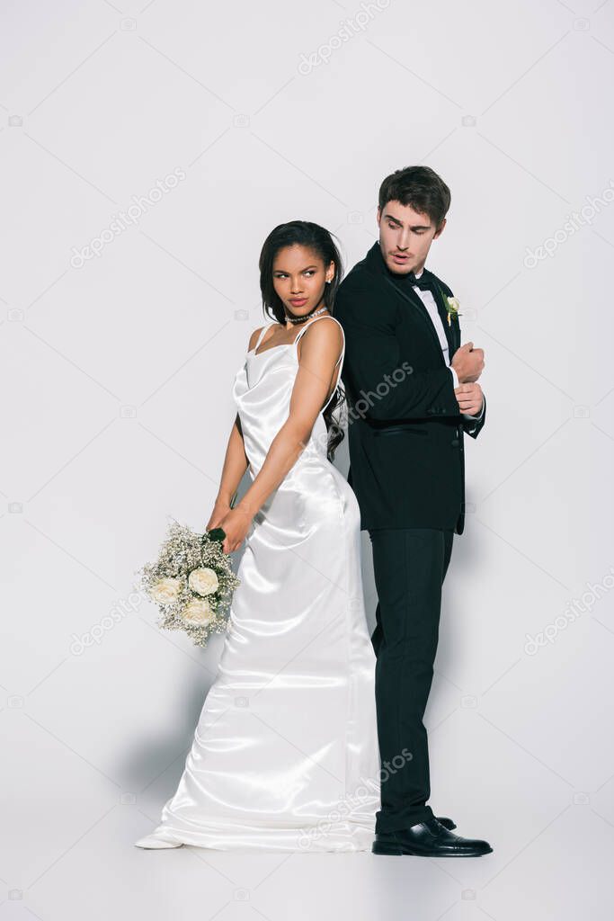 full length view of elegant interracial newlyweds standing back to back on white background